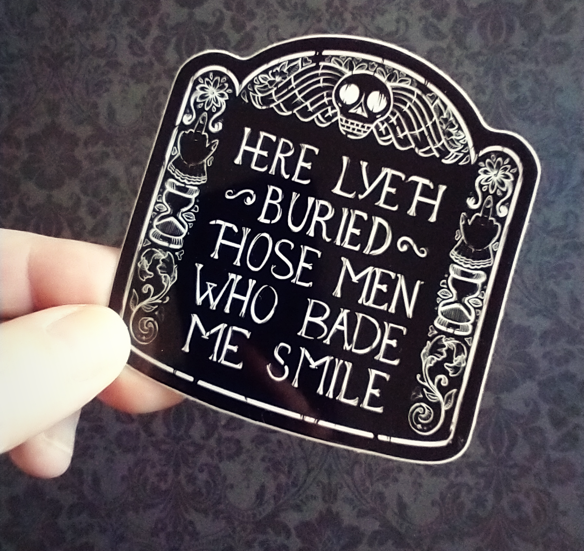 A hand holding a black and white sticker bearing an illustration of a headstone, which reads, "Here lyeth buried those men who bade me smile".