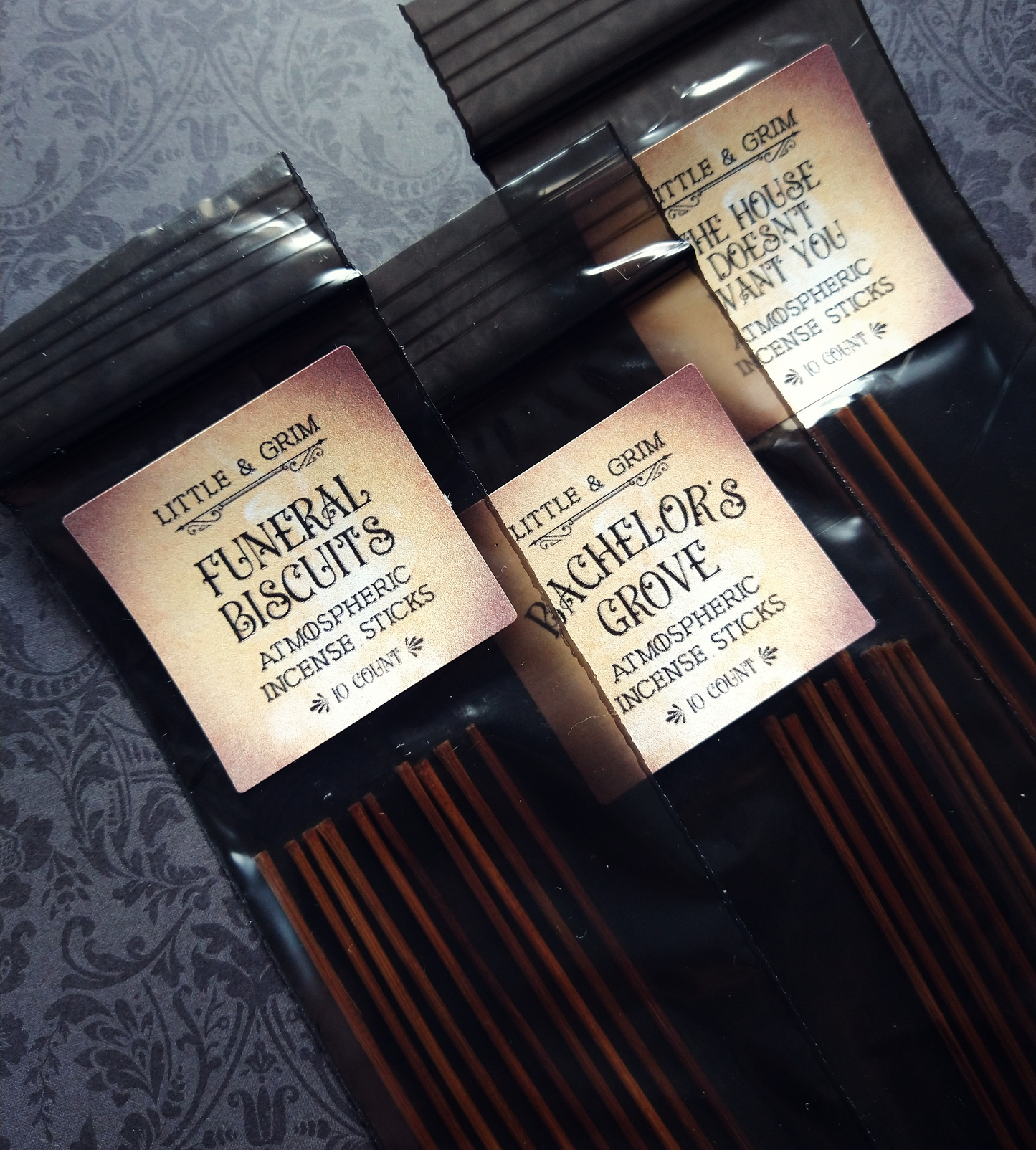 Three bags of Little and Grim incense, presented in black bags with labels bearing the scent names: "Funeral Biscuits", "Bachelor's Grove", and "The House Doesn't Want You."