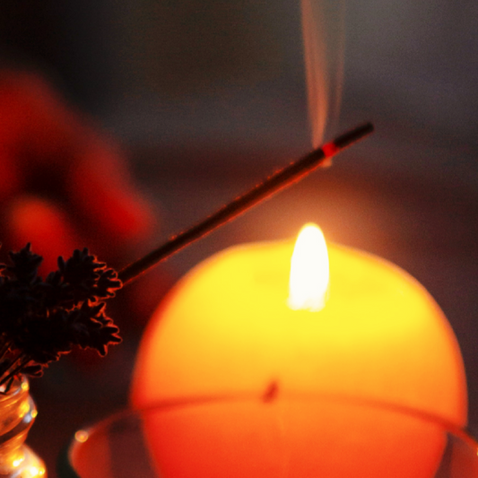 A close-up of a burning incense stick in front of a lit candle.