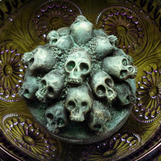 A Catacomb soap in an olive glass bowl. The soap features many small skulls piled together, and is slightly shimmery green in tone.